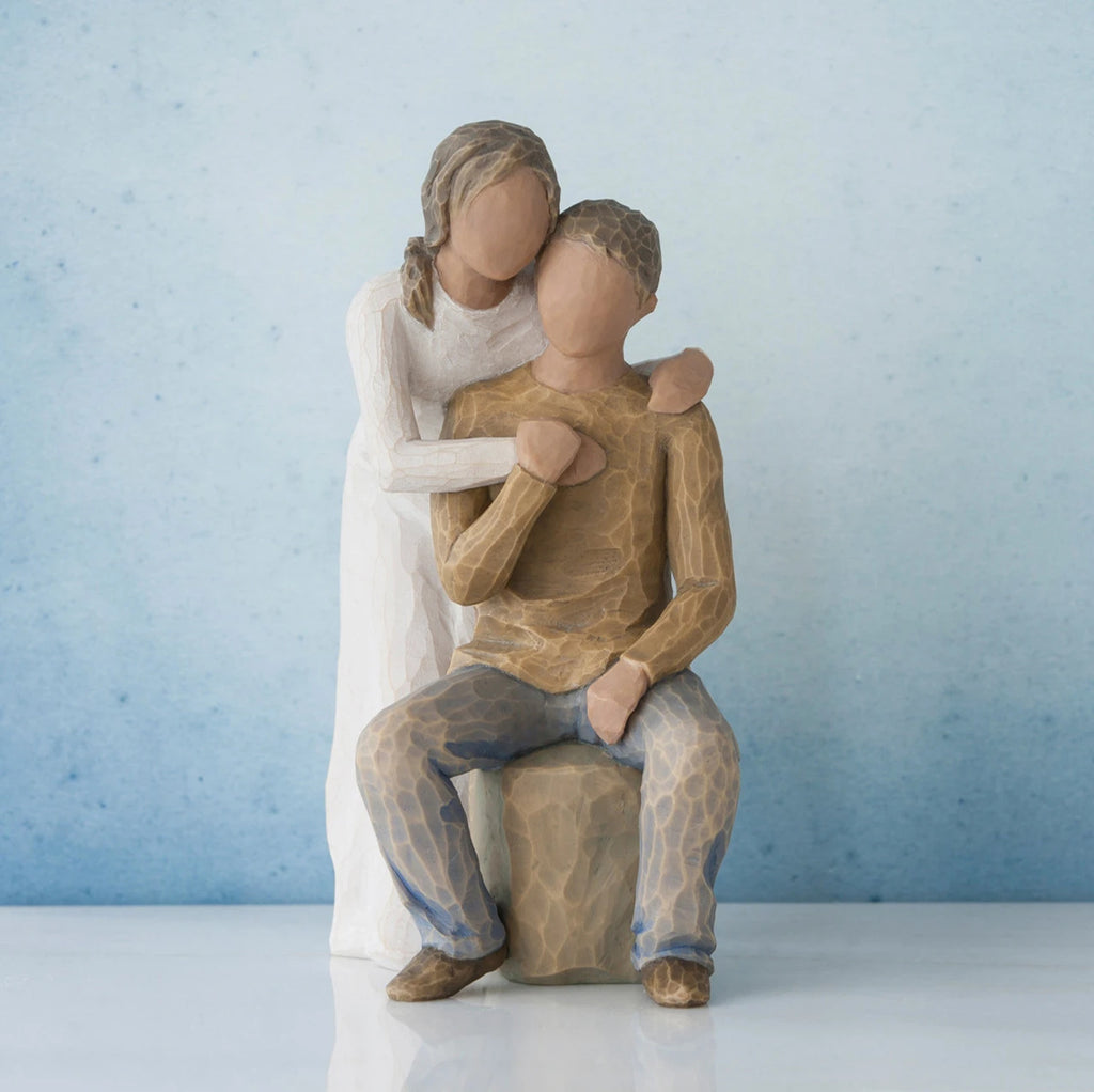 Man figurine sitting on rock wearing brown shirt and blue jeans with woman figurine in white dress with arms wrapped around him - blue background
