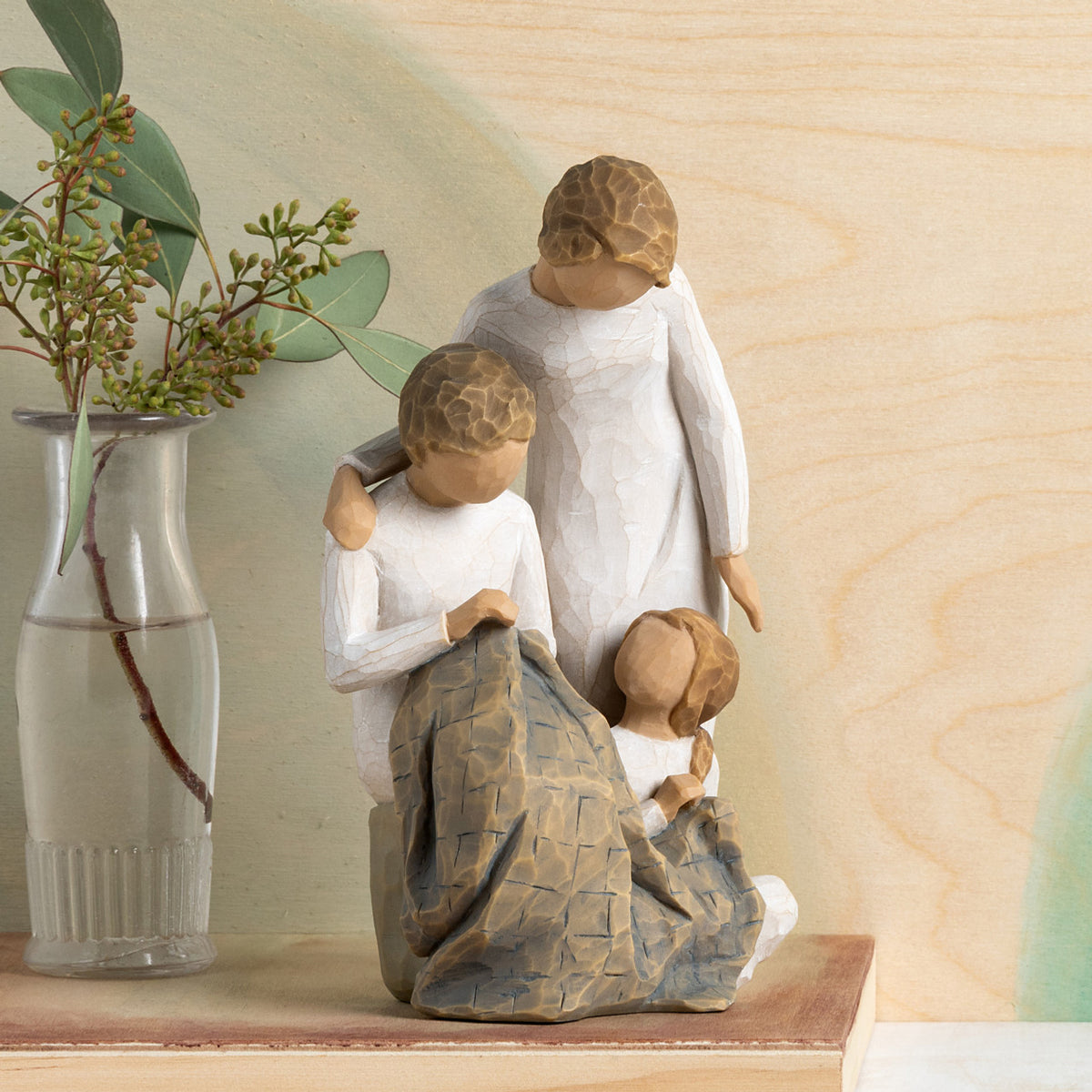 Willow Tree Generations, 7”h hand-painted resin figure of seated older woman sewing blue quilt on her lap; standing woman at her shoulder; seated young girl at her feet, all in neutral clothing, on table with green florals