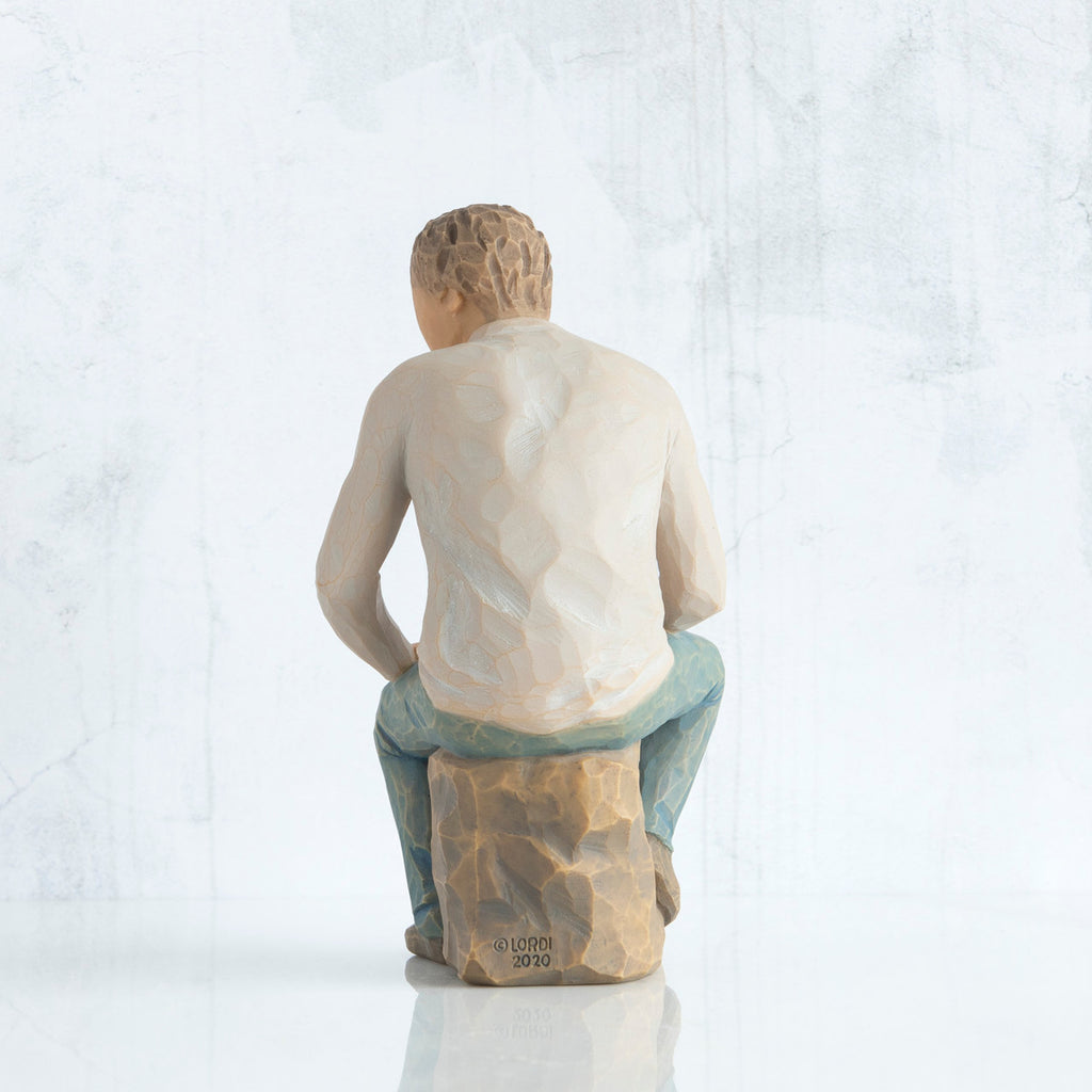 Back view, adult male figure in light gray shirt and blue jeans, seated on gray rock