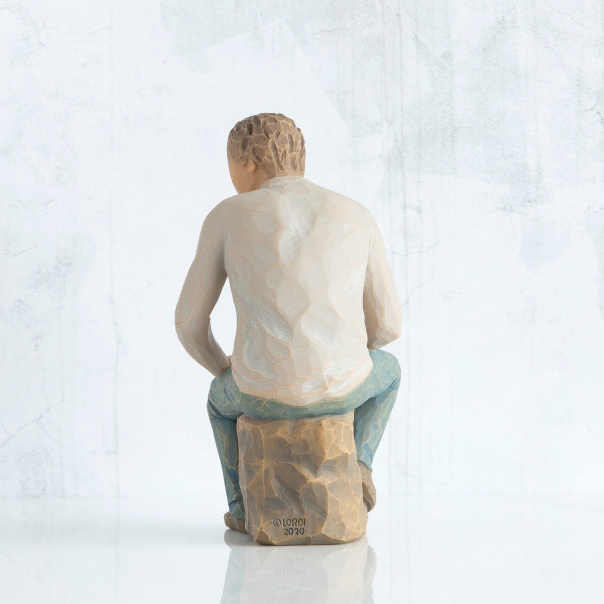 Back view, adult male figure in light gray shirt and blue jeans, seated on gray rock