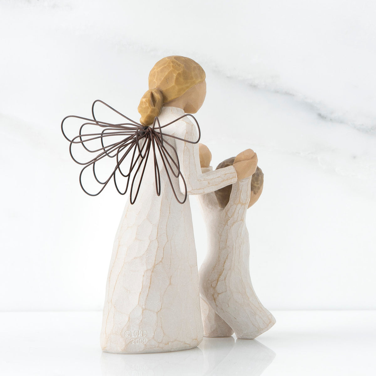 Blonde angel figurine in white dress, black wire wings, facing the back holding baby figurine's hands up
