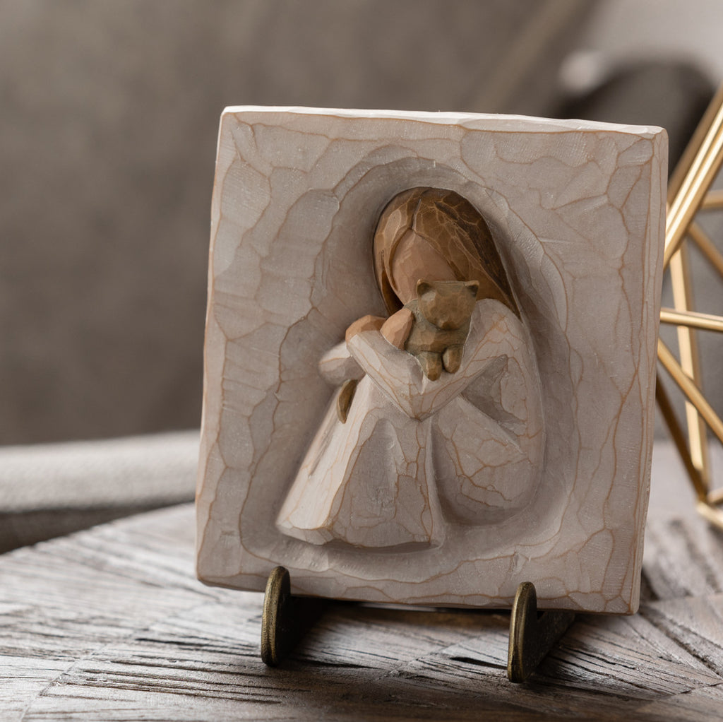 Willow Tree Comfort Plaque, 4”sq x .5”d hand-painted resin bas-relief carving of seated figure in cream dress, holding small gray cat. Plaque displayed on Plaque Stand on table