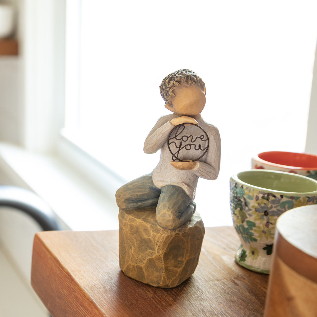 Willow Tree Love you too (lighter skin), 4.5”h hand-painted resin figure of young child in cream shirt and jeans, kneeling on rock, holding wire circle with ‘love you’ inside, on shelf by window